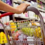 A imagem mostra um carrinho de supermercado sendo empurrado em frente a um corredor do mercado. O fundo está desfocado, mas há itens de limpeza. O carrinho está cheio, com uva, banana e outros itens.