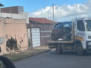 Imagem mostra, à esquerda, um muro amarelo com um buraco grande e irregular, com diversas rachaduras. À direita, um carro HB20 preto com a parte dianteira destruída, em cima de um guincho.