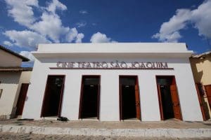 A imagem mostra uma construção vista por baixo. É uma casa térrea, branca. Há quatro portas de madeira, uma do lado da outra, e um letreiro acima delas diz "Cine Teatro São Joaquim".