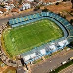 A imagem mostra um estádio visto de cima. É possível ver as marcações do campo no chão, e as arquibancadas azuis ocupam três dos lados do estádio.