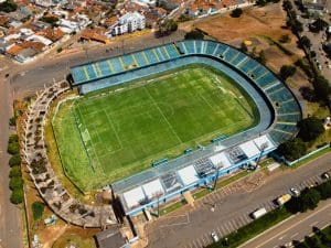 A imagem mostra um estádio visto de cima. É possível ver as marcações do campo no chão, e as arquibancadas azuis ocupam três dos lados do estádio.