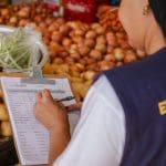 A imagem mostra uma mulher pelas costas. Ela está anotando coisas em uma prancheta, e está em frente a uma bancada de verduras.