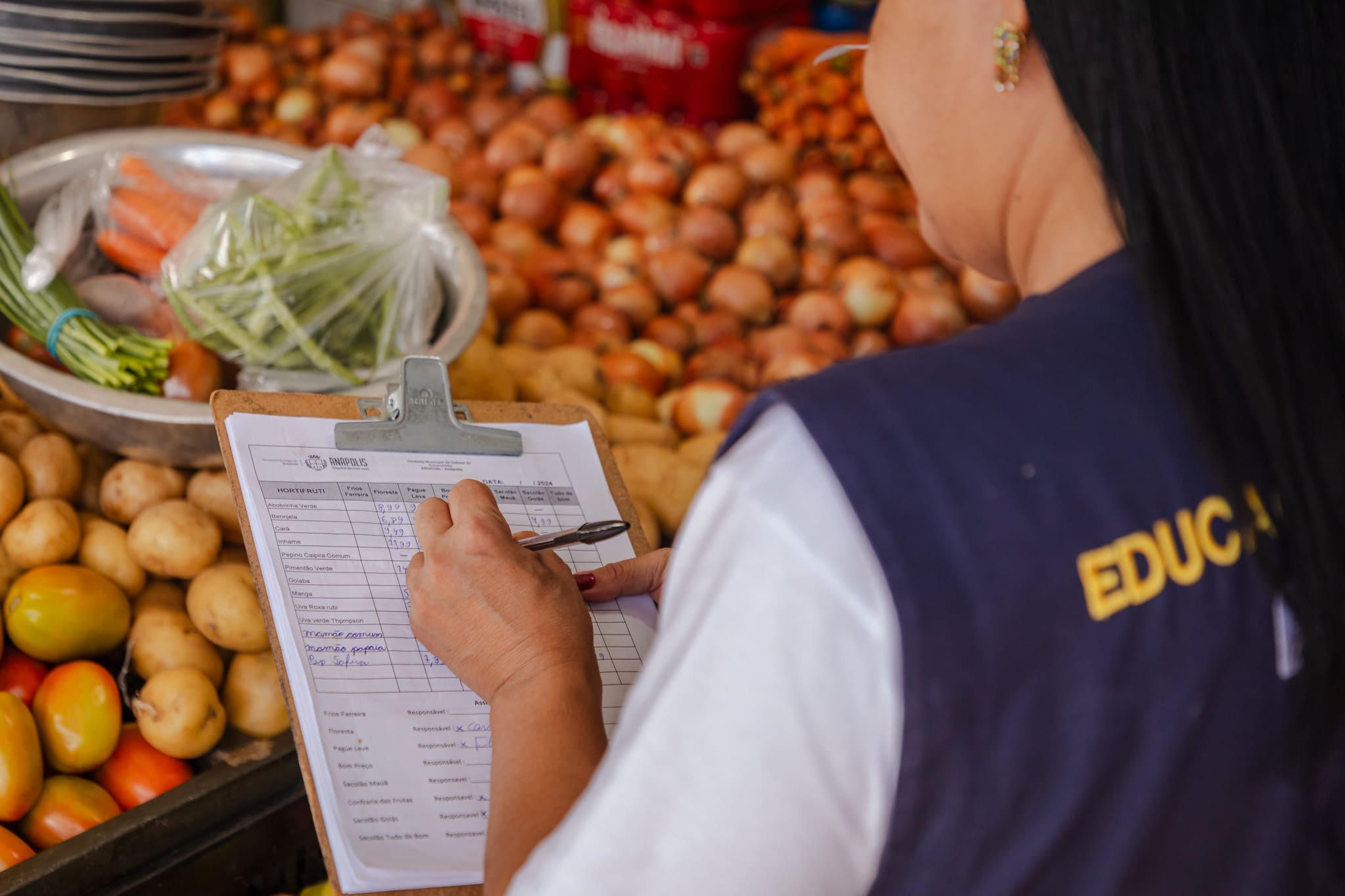 A imagem mostra uma mulher pelas costas. Ela está anotando coisas em uma prancheta, e está em frente a uma bancada de verduras.