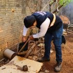 A imagem mostra um homem com um colete azul da Vigilância em Saúde. Ele está inclinado, mexendo em entulhos no chão, e procura por focos do mosquito da dengue.