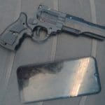 A imagem mostra um revólver (um simulacro de arma de fogo) e um aparelho celular, que tem a tela desligada. Ambos estão sobre uma superfície preta.