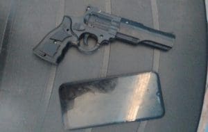 A imagem mostra um revólver (um simulacro de arma de fogo) e um aparelho celular, que tem a tela desligada. Ambos estão sobre uma superfície preta.