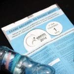 A imagem mostra uma prova de capa azul, uma garrafa de água de plástico, e uma caneta preta. A prova é do Enem 2014. O fundo é totalmente preto.