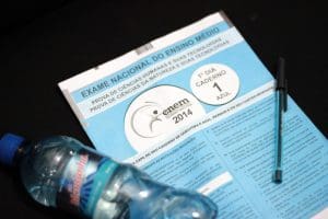 A imagem mostra uma prova de capa azul, uma garrafa de água de plástico, e uma caneta preta. A prova é do Enem 2014. O fundo é totalmente preto.