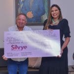 A imagem mostra Vilmar Mariano, um homem branco e baixo, e Silvye Alves, mulher branca, alta e de cabelos longos. Eles seguram um grande cheque simbólico que indica o valor de R$5,1 milhões.