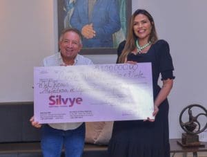 A imagem mostra Vilmar Mariano, um homem branco e baixo, e Silvye Alves, mulher branca, alta e de cabelos longos. Eles seguram um grande cheque simbólico que indica o valor de R$5,1 milhões.