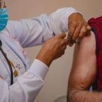 A imagem mostra duas pessoas. A primeira, à esquerda, é uma profissional da saúde que usa um jaleco branco, uma máscara azul e um óculos preto. Ela aplica uma vacina em um senhor, à direita, que usa uma camiseta vermelha com a manga erguida.
