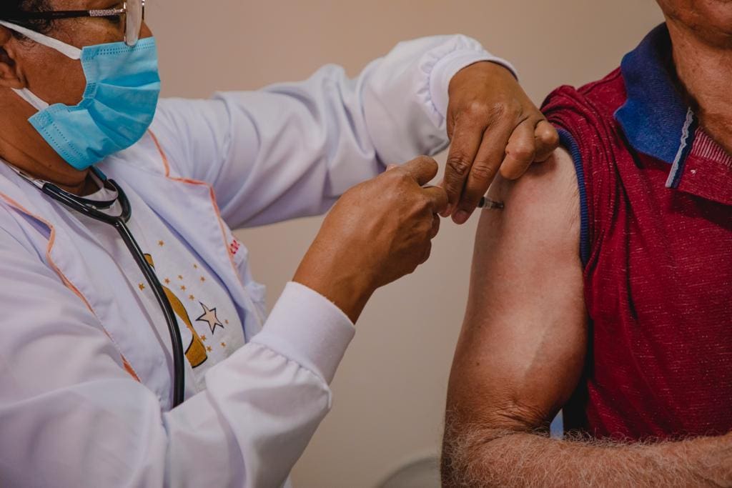 A imagem mostra duas pessoas. A primeira, à esquerda, é uma profissional da saúde que usa um jaleco branco, uma máscara azul e um óculos preto. Ela aplica uma vacina em um senhor, à direita, que usa uma camiseta vermelha com a manga erguida.