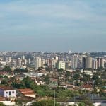 A imagem mostra uma vista aérea da cidade de Anápolis. A parte superior da imagem mostra o céu azul. Já a inferior contém casas, mais perto, e prédios, ao longe.