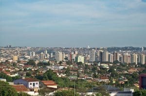 A imagem mostra uma vista aérea da cidade de Anápolis. A parte superior da imagem mostra o céu azul. Já a inferior contém casas, mais perto, e prédios, ao longe.