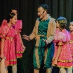 Um homem, vestido de verde, e quatro garotas (crianças) vestidas de rosa conversam em cima de um palco. Elas têm o cabelo trançado e ele usa um microfone. Estão encenando uma peça teatral, e o fundo é escuro.