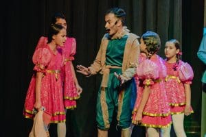 Um homem, vestido de verde, e quatro garotas (crianças) vestidas de rosa conversam em cima de um palco. Elas têm o cabelo trançado e ele usa um microfone. Estão encenando uma peça teatral, e o fundo é escuro.