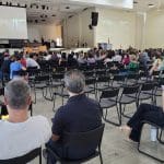 Diversos professores adultos, homens e mulheres, estão sentados em cadeiras pretas diante de um palco e uma tela.