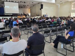 Diversos professores adultos, homens e mulheres, estão sentados em cadeiras pretas diante de um palco e uma tela.