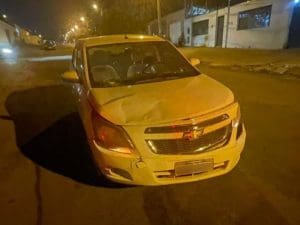 A imagem mostra a parte dianteira de um carro branco da marca Chevrolet. O capô está amassado, à esquerda, e o para-choque está fora do lugar, à direita. Acima do ponto em que o capô está amassado, o vidro está rachado. A foto foi tirada à noite.