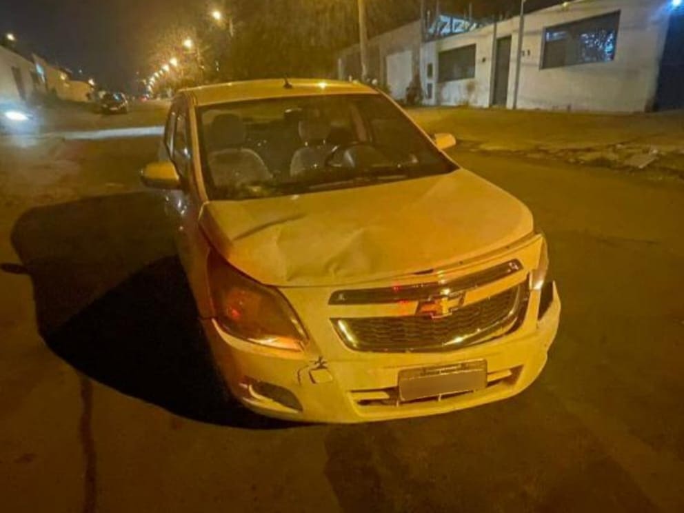 A imagem mostra a parte dianteira de um carro branco da marca Chevrolet. O capô está amassado, à esquerda, e o para-choque está fora do lugar, à direita. Acima do ponto em que o capô está amassado, o vidro está rachado. A foto foi tirada à noite.