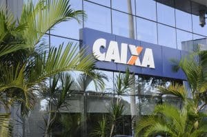 Imagem mostra a fachada de uma agência da Caixa Econômica Federal. A palavra "Caixa" está em letras brancas e laranjas sobre uma placa azul. Plantas cercam a fachada, formando uma moldura.