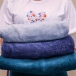 Uma pessoa de camiseta branca segura nas mãos três cobertores azuis, um sobre o outro. Não é possível ver seu rosto.