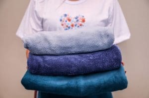 Uma pessoa de camiseta branca segura nas mãos três cobertores azuis, um sobre o outro. Não é possível ver seu rosto.