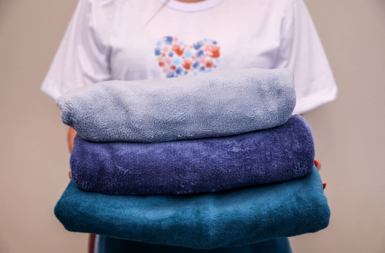 Uma pessoa de camiseta branca segura nas mãos três cobertores azuis, um sobre o outro. Não é possível ver seu rosto.