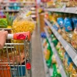 Alguém empurra um carrinho de supermercado ao lado de prateleiras cheias de comida.
