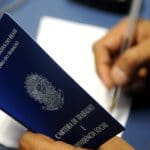 O primeiro plano da imagem mostra uma mão segurando uma carteira de trabalho azul, que está aberta. No fundo desfocado, a mão direita usa uma caneta para escrever em uma folha em branco. A mesa embaixo da folha é azul.