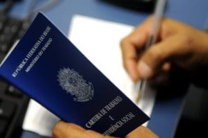 O primeiro plano da imagem mostra uma mão segurando uma carteira de trabalho azul, que está aberta. No fundo desfocado, a mão direita usa uma caneta para escrever em uma folha em branco. A mesa embaixo da folha é azul.