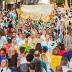 Várias pessoas preenchem toda a imagem. No centro, um bispo está debaixo de uma tenda carregada por fiéis e levanta uma imagem sagrada.