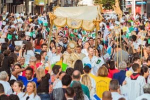 Várias pessoas preenchem toda a imagem. No centro, um bispo está debaixo de uma tenda carregada por fiéis e levanta uma imagem sagrada.