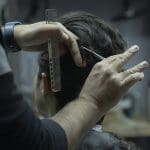 Alguém de cabelo curto e castanho tem o cabelo cortado por um profissional. O cabeleireiro veste preto, tem uma tesoura e um pente nas mãos.