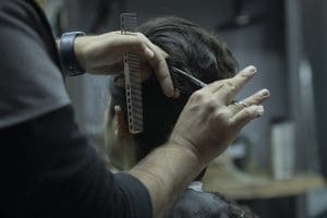 Alguém de cabelo curto e castanho tem o cabelo cortado por um profissional. O cabeleireiro veste preto, tem uma tesoura e um pente nas mãos.