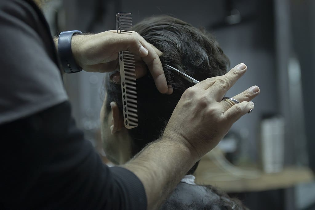 Alguém de cabelo curto e castanho tem o cabelo cortado por um profissional. O cabeleireiro veste preto, tem uma tesoura e um pente nas mãos.