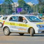 Um carro branco com uma faixa amarela escrito "autoescola" está parado em uma rua, visto de lado. Outros carros de autoescola estão atrás.
