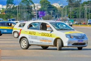 Um carro branco com uma faixa amarela escrito "autoescola" está parado em uma rua, visto de lado. Outros carros de autoescola estão atrás.
