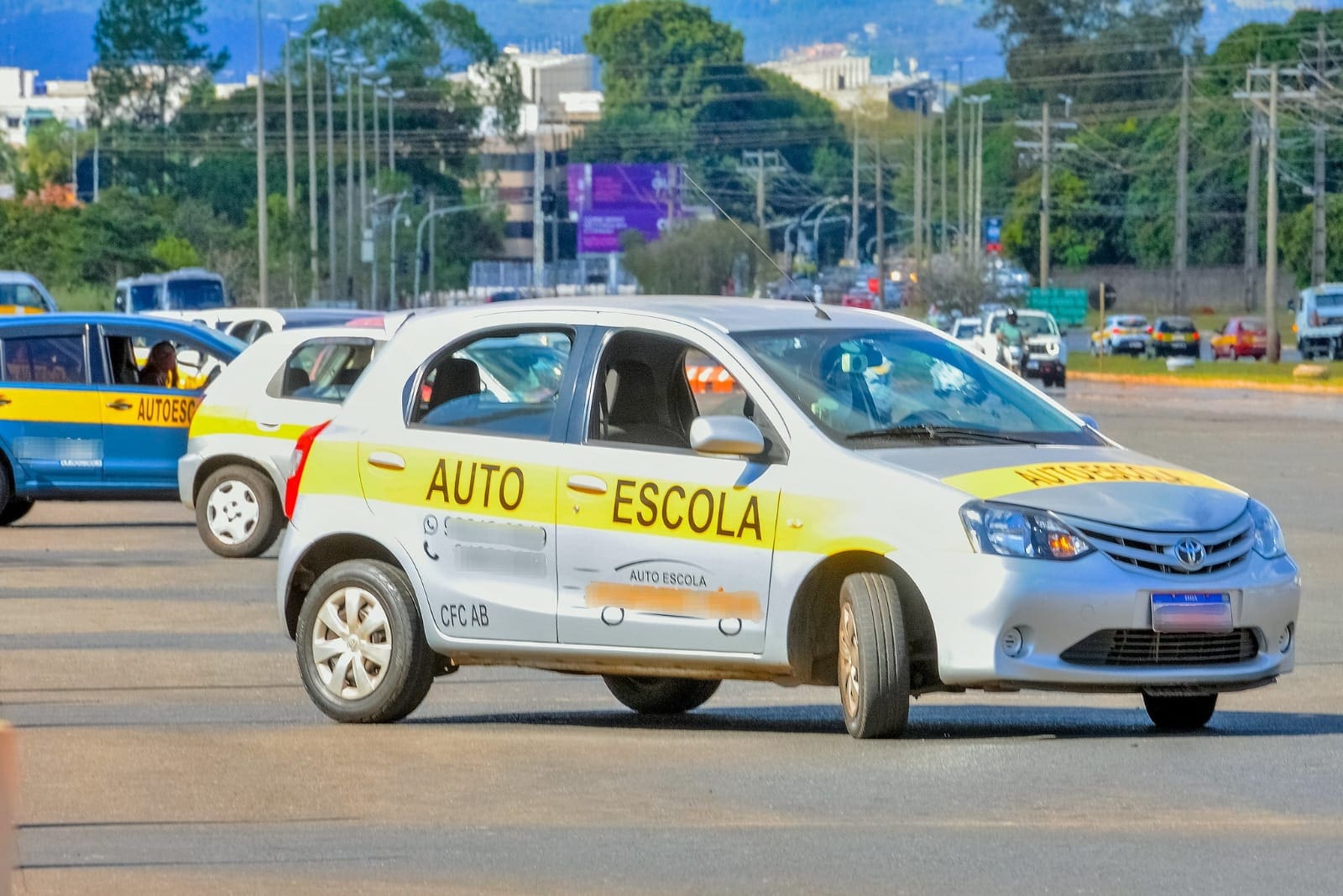 Um carro branco com uma faixa amarela escrito "autoescola" está parado em uma rua, visto de lado. Outros carros de autoescola estão atrás.
