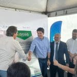 Dois homens brancos apertam as mãos em frente a uma placa do Governo de Goiás, que diz "ampliação do sistema de abastecimento de água em Anápolis). O da direita usa camisa azul claro, e o da esquerda veste uma camisa branca. Outros dois homens estão à direita.