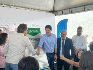Dois homens brancos apertam as mãos em frente a uma placa do Governo de Goiás, que diz "ampliação do sistema de abastecimento de água em Anápolis). O da direita usa camisa azul claro, e o da esquerda veste uma camisa branca. Outros dois homens estão à direita.