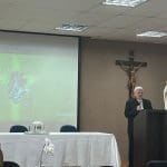 À direita, um bispo está de pé, atrás de púlpito e com um microfone na mão. Há uma cruz na parede às suas costas. À esquerda, um projetor mostra as palavras "58º Dia Mundial das Comunicações Sociais".