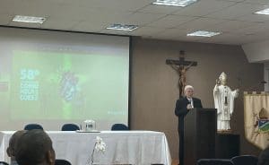 À direita, um bispo está de pé, atrás de púlpito e com um microfone na mão. Há uma cruz na parede às suas costas. À esquerda, um projetor mostra as palavras "58º Dia Mundial das Comunicações Sociais".