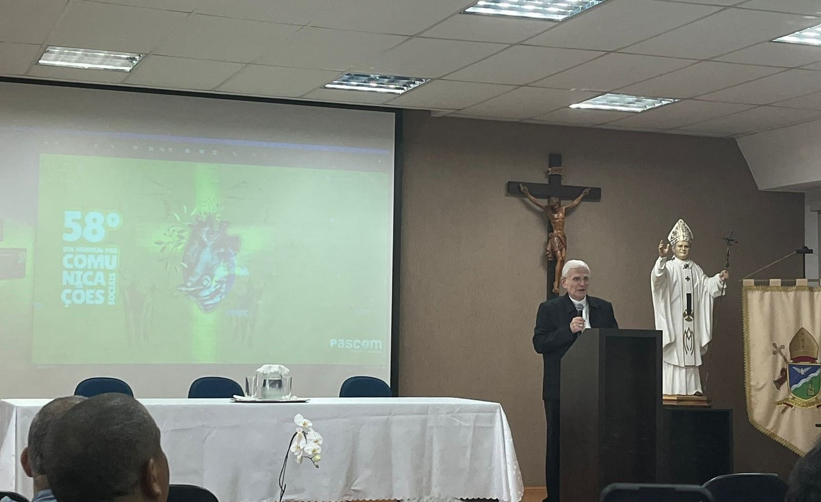 À direita, um bispo está de pé, atrás de púlpito e com um microfone na mão. Há uma cruz na parede às suas costas. À esquerda, um projetor mostra as palavras "58º Dia Mundial das Comunicações Sociais".