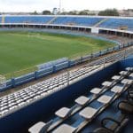 Um estádio é visto por dentro, a partir das arquibancadas. É possível ver metade do gramado, com um gol, e parte das arquibancadas.