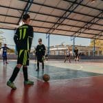 Há um garoto de uniforme preto com detalhes verdes neon., que joga futsal com outros meninos. Ele está de costas, se preparando para chutar uma bola do escanteio de uma quadra. A camisa diz "futsal" e "11" em letras verdes.