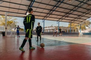 Há um garoto de uniforme preto com detalhes verdes neon., que joga futsal com outros meninos. Ele está de costas, se preparando para chutar uma bola do escanteio de uma quadra. A camisa diz "futsal" e "11" em letras verdes.