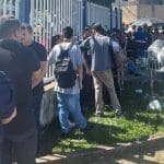 A imagem mostra uma fila de pessoas ao lado de uma grade branca. Elas estão de costas e algumas pessoas conversam.