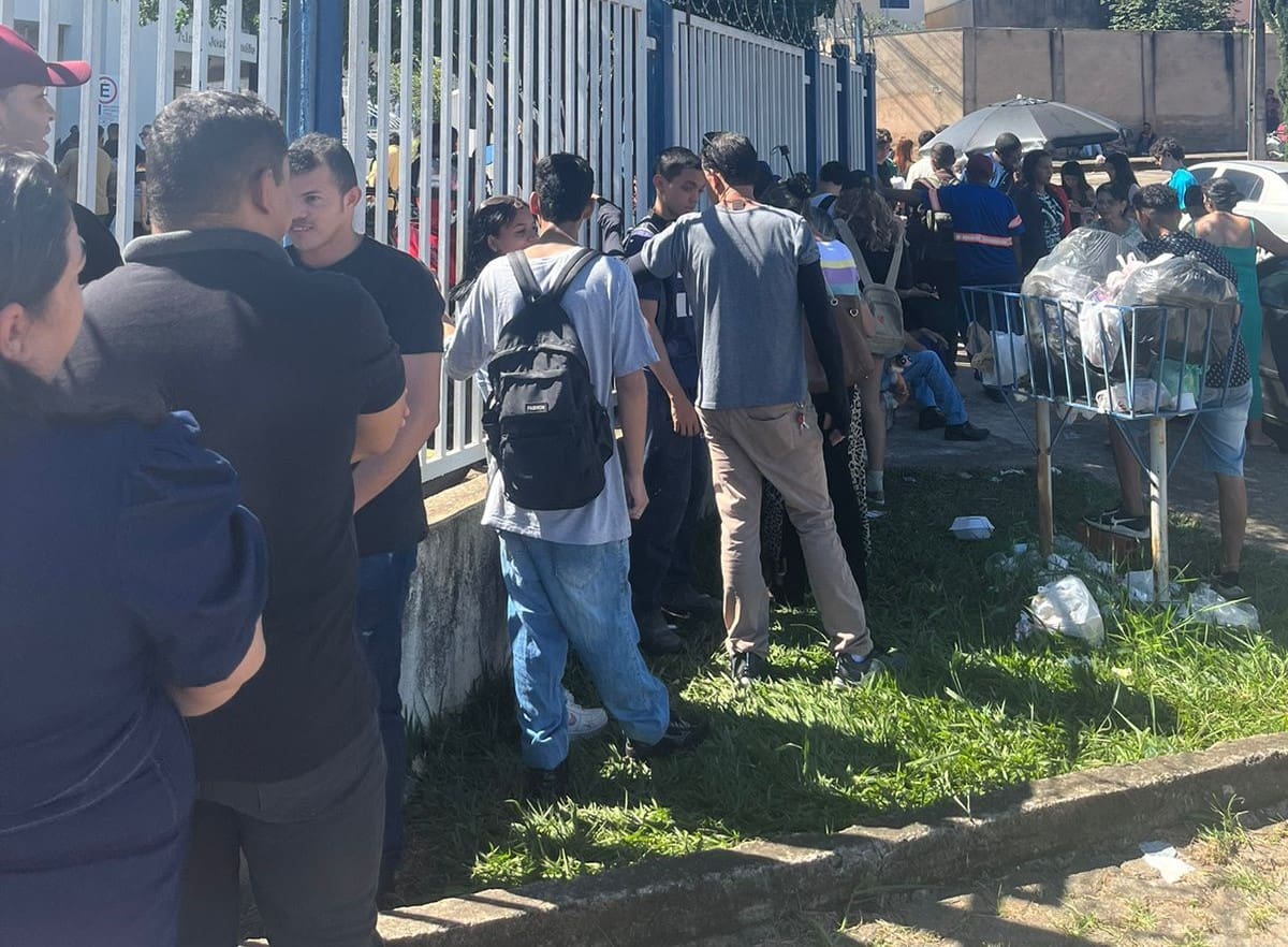 A imagem mostra uma fila de pessoas ao lado de uma grade branca. Elas estão de costas e algumas pessoas conversam.