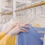 A imagem mostra os braços de uma mulher branca mexendo em cabides transparentes em uma arara de roupas. As mangas da mulher são amarelas, e no cabide há um casaco azul.